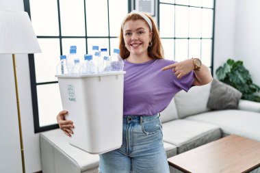 Geri dönüşüm sepetini elinde plastik şişelerle tutan genç, kızıl saçlı bir kadın. Kendine güvenen, gülümseyen, kendini gururlu ve mutlu gösteren bir kadın.. 