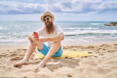 Genç kızıl saçlı adam kumsalda havluya oturmuş akıllı telefon kullanıyor..