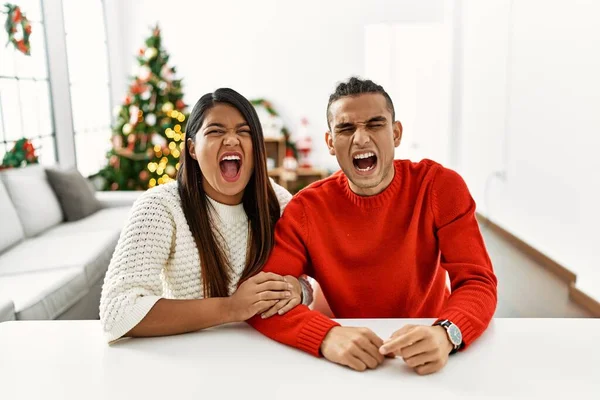 Young Latin Couple Sitting Table Christmas Tree Angry Mad Screaming — Zdjęcie stockowe