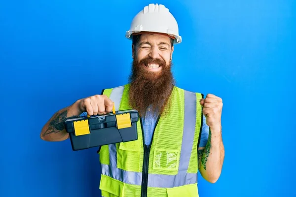 Redhead man with long beard wearing safety helmet and reflective jacket holding toolbox screaming proud, celebrating victory and success very excited with raised arm
