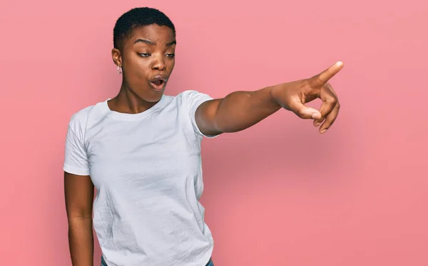 若いアフリカ系アメリカ人の女性がカジュアルな白いTシャツを着て 指で前に驚いて 口を開けて驚いた表情 正面に何か — ストック写真