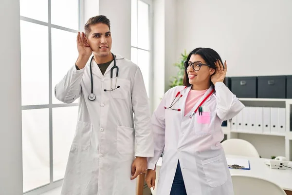 Young doctors wearing uniform and stethoscope at the clinic smiling with hand over ear listening and hearing to rumor or gossip. deafness concept.