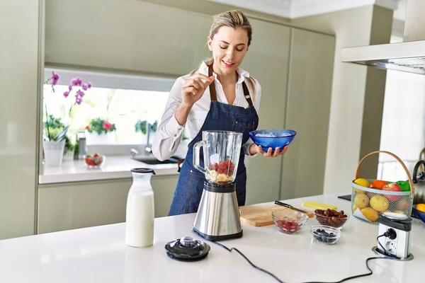 Genç sarışın kadın mutfaktaki blendere buz dökerek gülümsüyor.