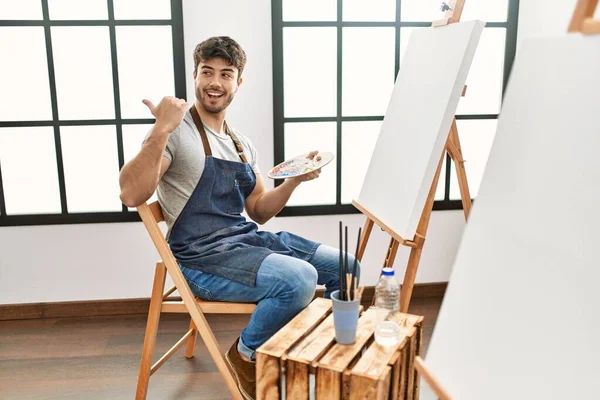 Young Hispanic Man Painting Art Studio Pointing Thumb Side Smiling — 图库照片