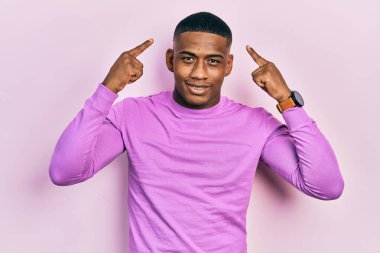 Genç siyah bir adam pembe bir süveter giyip gülümsüyor ve iki eliyle kafasını işaret ediyor. Harika bir fikir ya da düşünce, iyi bir hafıza. 