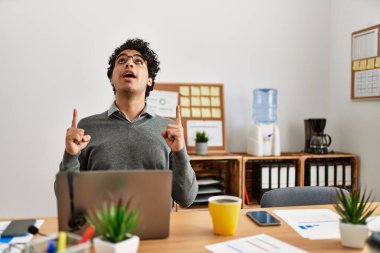 Young hispanic man wearing business style sitting on desk at office amazed and surprised looking up and pointing with fingers and raised arms. 