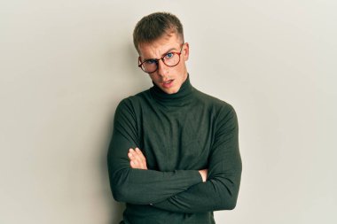 Kollarını kavuşturmuş gözlük takan genç beyaz bir adam. Şok olmuş yüz ifadesi. Şüpheci ve alaycı görünüyor. 