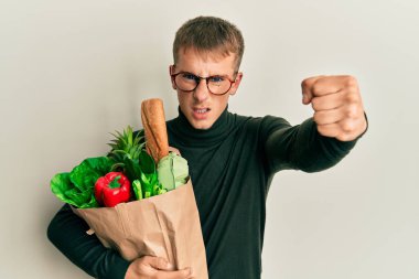 Kafkasyalı genç bir adam elinde alışveriş torbalarıyla sinirlenmiş ve sinirlenmiş bir şekilde bağırıyor. Öfkeyle bağırıyor ve el kaldırıyor. 