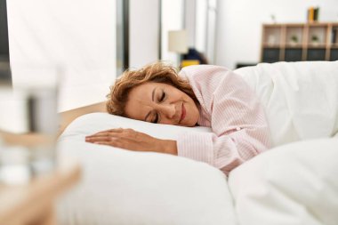 Orta yaşlı beyaz kadın yatak odasında yatakta yatıyor..