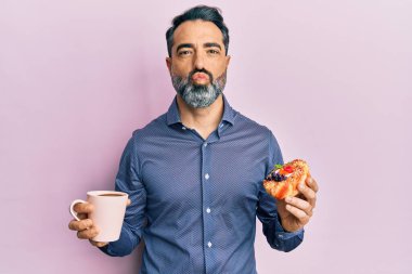 Orta yaşlı, sakallı, kır saçlı kahve içen ve kameraya bakarak pasta yiyen bir adam. Güzel ve seksi. aşk ifadesi. 