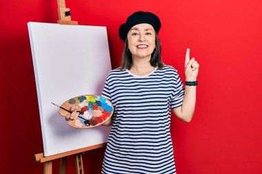 Orta yaşlı İspanyol bir kadın elinde paletle ayakta dikiliyor. Yüzünde kocaman bir gülümsemeyle resim sehpası duruyor. Elleriyle kameraya bakıyor.. 