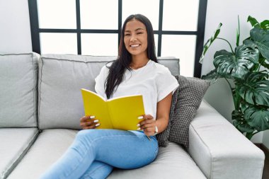 Genç Latin kadın gülümsüyor evde kendine güvenen kitap okuyor.