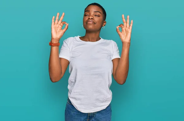 カジュアルな白いTシャツを着た若いアフリカ系アメリカ人女性がリラックスして笑顔で目を閉じて指で瞑想ジェスチャーをしています ヨガのコンセプト — ストック写真