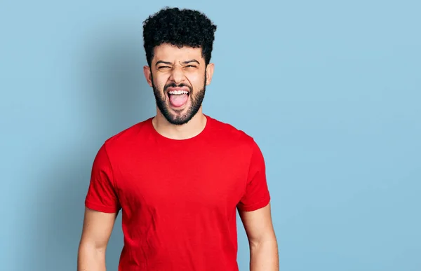 年轻的阿拉伯男子留着胡子 穿着休闲的红色T恤 带着滑稽的表情伸出舌头 情感概念 — 图库照片