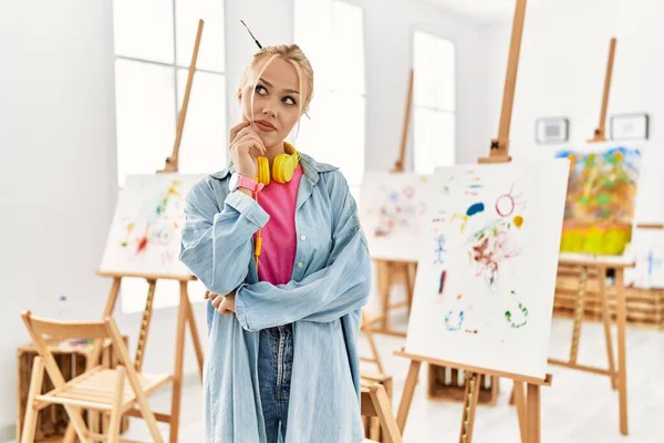 Young caucasian girl at art studio with hand on chin thinking about question, pensive expression. smiling with thoughtful face. doubt concept.