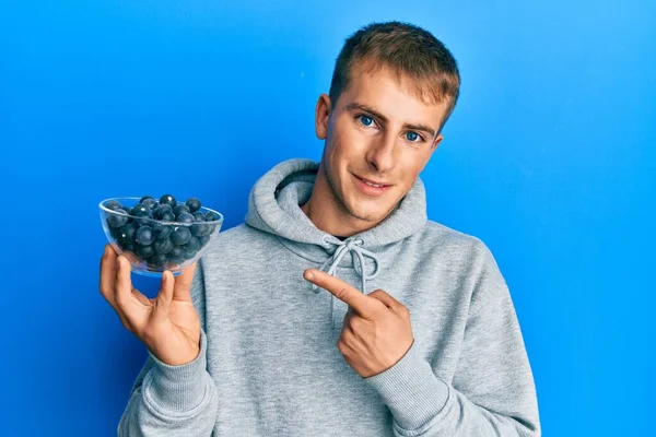Young Caucasian Man Holding Bowl Blueberries Smiling Happy Pointing Hand — стоковое фото