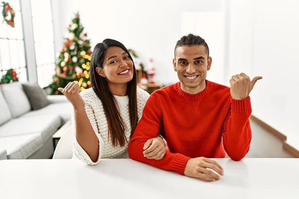 Young Latin Couple Sitting Table Christmas Tree Pointing Back Hand — Stockfoto