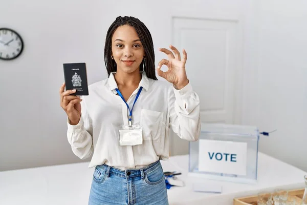 Young african american woman at political campaign election holding canada passport doing ok sign with fingers, smiling friendly gesturing excellent symbol