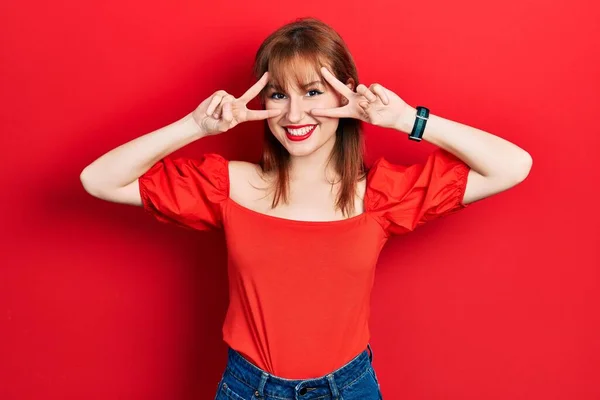 顔の上に指で平和の象徴を行うカジュアルな赤いTシャツを着て若い女性をRedhead 勝利を示す陽気な笑顔 — ストック写真