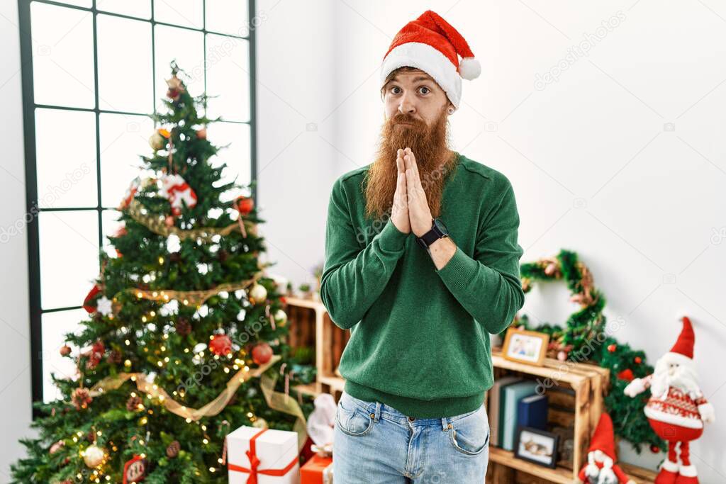 Redhead man with long beard wearing christmas hat by christmas tree praying with hands together asking for forgiveness smiling confident. 