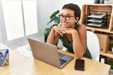 Ofiste çalışırken laptopunu kullandığından emin bir şekilde gülümseyen genç İspanyol kadın.