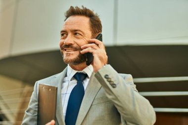 Orta yaşlı işadamı gülümsüyor. Şehirdeki akıllı telefondan konuşuyor..