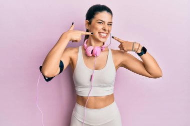 Spor kıyafetleri giyen, kulaklık takan, gülümseyen, parmaklarıyla ve ağzıyla işaret eden İspanyol bir kadın. diş sağlığı kavramı. 