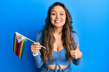 Zimbabwe bayrağı tutan genç İspanyol kız mutlu ve pozitif gülümsüyor, başparmağıyla mükemmel ve onay işareti yapıyor. 