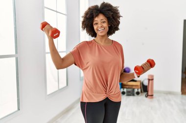 Genç Afrikalı Amerikalı kadın gülümsüyor spor merkezinde halterleri kullanarak kendine güven eğitimi alıyor