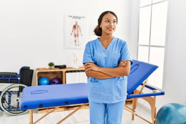 Klinikte kollarını kavuşturmuş duran fizyoterapist üniforması giyen genç Latin kadın.