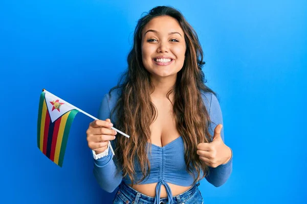 Zimbabwe bayrağı tutan genç İspanyol kız mutlu ve pozitif gülümsüyor, başparmağıyla mükemmel ve onay işareti yapıyor. 