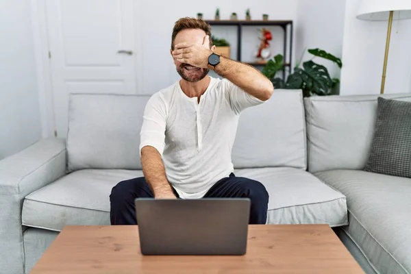 Middle age man using laptop at home smiling and laughing with hand on face covering eyes for surprise. blind concept.