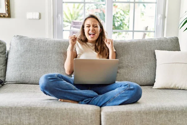 腕を上げて成功のために興奮して自宅でコンピュータのラップトップを使用してソファの上に座っている美しい若いブルネットの女性と目は勝利笑顔を祝う閉じました 勝者のコンセプト — ストック写真