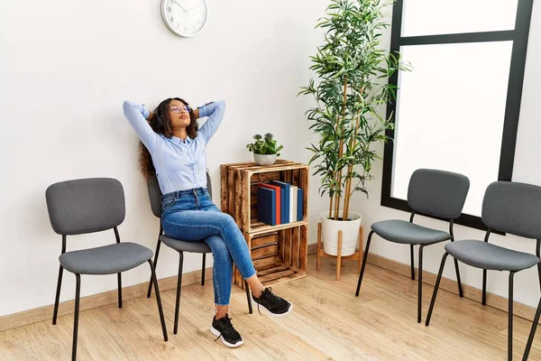 Genç Latin kadın ellerini başının üstüne koyup bekleme odasında müzik dinliyor.