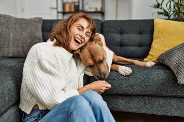 Beyaz kadın gülümsüyor. Kendine güvenen, kucaklayan köpek evde yerde oturuyor.