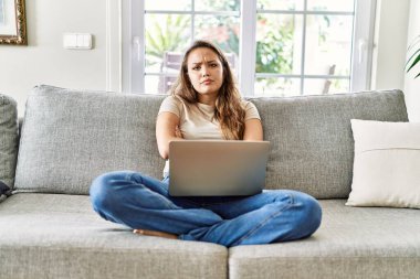 Kanepede oturan genç, güzel esmer kadın evde bilgisayar laptopu kullanıyor şüpheci ve gergin, yüzünde çapraz kollu tasvip etmeyen bir ifade. negatif kişi. 