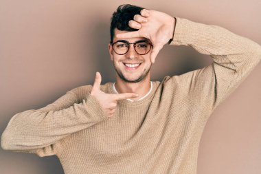 Gündelik kıyafetler ve gözlük takan genç İspanyol adam gülümsüyor ve mutlu yüzlü eller ve parmaklarla çerçeve yapıyor. yaratıcılık ve fotoğrafçılık kavramı. 