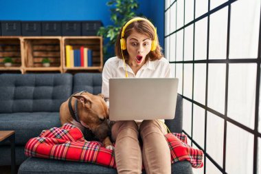 Kanepede dizüstü bilgisayar kullanan genç ve güzel bir kadın korkmuş ve ağzı açık bir şekilde şaşırmış bir şekilde oturuyor. 