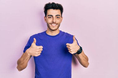 Gündelik tişört giyen genç İspanyol adam. Başarı işareti ile el hareketi yapıyor. Gülümsüyor ve mutlu. Neşeli ifade ve kazanan jest. 