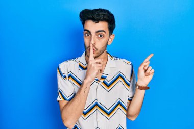 Gündelik giysiler giyen genç İspanyol adam sessiz olmak istiyor. Parmakları dudaklarında, elleri yandan işaret ediyor. sessizlik ve gizli konsept. 