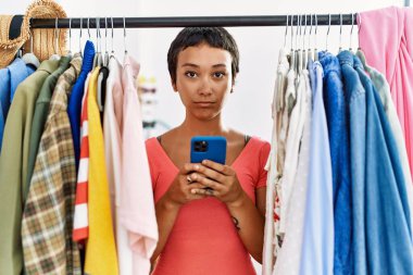 Kısa saçlı genç İspanyol kadın elbise rafında kıyafet arıyor. Akıllı telefonu kullanıyor. Düşünme tavrı takınıyor ve ayık bir yüz ifadesi takınıyor. 