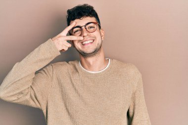 Gündelik kıyafetler giyen ve gözlük takan genç İspanyol adam barış sembolü yapıyor. Yüzünde parmaklar, gülümseyerek zaferi gösteriyor. 