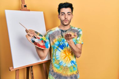 Genç İspanyol adam paletle resim çiziyor, resim sehpası ayakta kameraya bakıyor, havada elle öpücük savuruyor, güzel ve seksi. aşk ifadesi. 