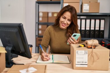 Genç Latin iş kadını akıllı telefon kullanıyor ve ofiste kitap yazıyor.