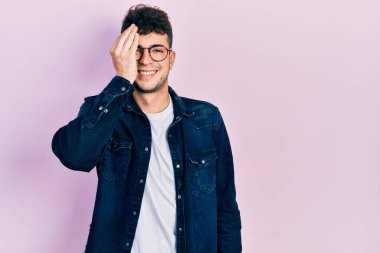 Gündelik kıyafetler ve gözlük takan genç İspanyol bir adam bir gözünü eliyle kapatıyor, yüzünde kendine güvenen bir gülümseme ve sürpriz bir duygu var.. 