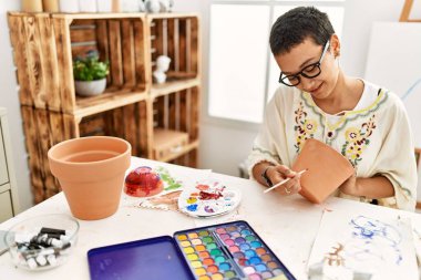 Young hispanic woman smiling confident painting clay ceramic at art studio clipart