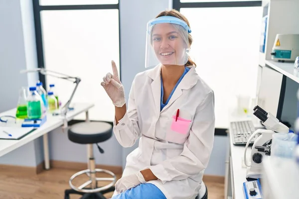 Young blonde woman working at scientist laboratory wearing face mask smiling with an idea or question pointing finger up with happy face, number one
