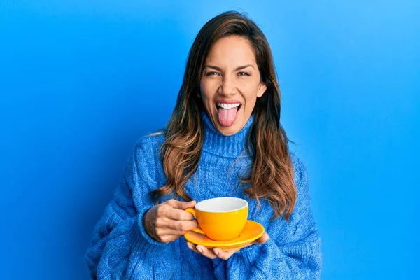 Young latin woman drinking a cup of coffee sticking tongue out happy with funny expression.