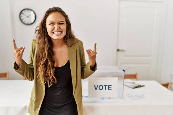 Beautiful hispanic woman standing at political campaign room shouting with crazy expression doing rock symbol with hands up. music star. heavy concept.