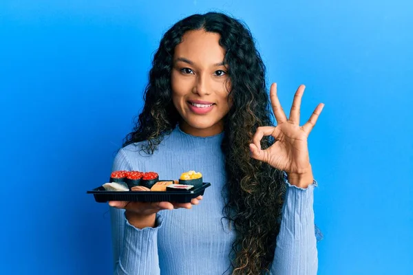 Bir tabak suşi gösteren genç Latin kadın parmaklarıyla işaret veriyor, gülümseyerek mükemmel bir sembol oluşturuyor. 